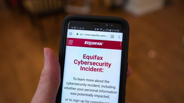 Gros plan de la main d'un homme tenant un téléphone portable ouvert sur le site Web du bureau de crédit Equifax, avec texte sur le site en cours de lecture 
