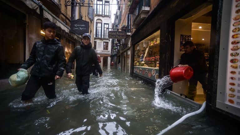 Un homme draine et tire de l'eau d'un magasin de souvenirs pendant que des adolescents marchent dans une ruelle inondée