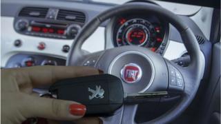 Une femme détient une clé de voiture Peugeot devant un volant Fiat