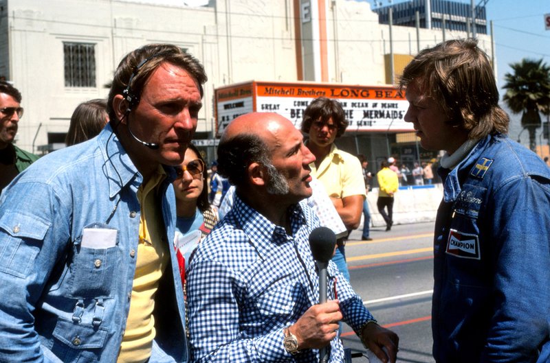 Dan Gurney, consultant pour le GPALB, se tient prêt comme ami et ancien rival Stirling Moss interviewe Ronnie Peterson avant la course de 1976. 